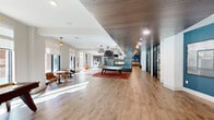 This expansive single-floor amenity space is thoughtfully designed to foster community and comfort, offering a generous open-plan layout finished with warm, light-toned hardwood flooring throughout. The space is flooded with natural light from floor-to-ceiling windows and large glass doors that frame views of the surrounding exterior, creating a bright and welcoming atmosphere.

The living and lounge area serves as the social heart of the space, anchored by a bold, tile-clad double-sided fireplace and a striking pool table set atop a vibrant patterned area rug, all beneath a series of oversized pendant lights with a rich wood-panel ceiling detail overhead. Adjacent to this, a dedicated game area features a shuffleboard table bathed in sunlight from large windows.

A built-in entertainment wall with a dark walnut finish houses a large flat-screen TV and is complemented by unique wall art, creating a relaxed lounge atmosphere. The dining area provides casual seating with bistro-style tables, while the kitchen supports the space's entertaining functions. A well-organized mailroom with built-in black metal mailboxes and a clean white countertop serves practical resident needs. Throughout, the design balances contemporary aesthetics with functional, community-oriented living.