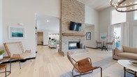 This thoughtfully designed single-floor amenity space offers an expansive, open-concept layout that seamlessly connects multiple functional zones. At the heart of the space stands a dramatic floor-to-ceiling brick fireplace with a reclaimed wood mantel and a mounted flat-screen TV, anchoring the main lounge area with warmth and visual impact. Soaring ceilings with wood-paneled accents amplify the sense of volume, while light wide-plank hardwood flooring flows consistently throughout, unifying each area.

The lounge features generous seating arrangements, including leather chairs and neutral sofas, creating inviting gathering spots. Large glass garage-style doors in the adjoining area flood the space with natural light and open to exterior views, effortlessly blending indoor and outdoor living. High-top seating near these doors adds a casual, social atmosphere.

The fully equipped kitchen is a standout feature, finished with sage green cabinetry, a textured white tile backsplash, and a waterfall-edge quartz island with bar seating. Stainless steel appliances, double wall ovens, and pendant lighting complete the functional yet stylish space.

A dining area accommodates group gatherings around a rustic farmhouse table. A display shelving unit and thoughtfully curated décor throughout contribute to the space's cohesive, contemporary coastal aesthetic.
