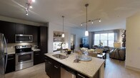 This single-floor space showcases a thoughtfully designed open-concept layout that seamlessly connects the kitchen, dining, and living areas. The kitchen serves as a central focal point, anchored by a generously sized island topped with light-toned granite countertops, an undermount sink, and elegant pendant lighting hanging overhead. Dark espresso cabinetry provides a striking contrast against the lighter surfaces, lending the space a contemporary, upscale aesthetic. Stainless steel appliances, including a built-in microwave and dishwasher, contribute to the kitchen's polished, modern character.

A dedicated nook with built-in shelving adds a decorative touch between the kitchen and living areas, offering display space for plants and décor. The living area extends naturally from the kitchen, benefiting from large windows that invite abundant natural light and offer views of the outdoors. A track lighting system supplements the pendant fixtures, ensuring the space is well-lit throughout. Light hardwood-style flooring runs throughout, tying the open floor plan together cohesively. The overall ambiance is sophisticated yet welcoming, balancing functional design with refined, contemporary finishes.