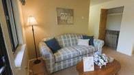 This single-floor space offers a warm and inviting atmosphere thoughtfully arranged for comfort. The living area serves as the heart of the space, centered around a plush plaid sofa in soft blue and white tones, accented with coordinating throw pillows that add a touch of casual elegance. A classic floor lamp with a warm-toned shade stands nearby, providing soft ambient lighting that enhances the room's cozy feel. The walls are finished in a gentle, neutral warm beige, creating a calm and restful backdrop throughout. A framed floral artwork hangs above the sofa, adding a decorative touch that complements the space's gentle color palette. A small round wooden side table holds a fresh floral arrangement, introducing a natural, cheerful element to the room. The carpeted flooring adds warmth underfoot and contributes to the overall sense of comfort. The layout is practical and uncluttered, allowing for easy movement through the space. The overall ambiance is quiet and residential, making it well-suited for relaxed living or a comfortable retreat. The thoughtful furnishings and soft color choices come together to create a space that feels both functional and welcoming.