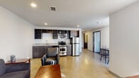 This single-floor unit presents a thoughtfully designed open-concept layout that seamlessly integrates the kitchen, dining, and living areas into one cohesive space. The kitchen is well-appointed with a full suite of stainless steel appliances, including a gas range, over-the-range microwave, dishwasher, and refrigerator. Dark espresso cabinetry with open upper shelving lines the kitchen wall, complemented by granite-style countertops and a single-basin sink. Polished concrete flooring extends throughout the main living area, lending a clean, contemporary aesthetic.

The living space accommodates both a seating area and dining setup, with natural light filtering in through large windows fitted with vertical blinds. A sliding glass-paneled interior door leads to the bedroom, which features plush carpet tile flooring, a dedicated desk workspace, and floor-to-ceiling windows offering outdoor views of the surrounding cityscape.

The bathroom is finished with a granite-topped vanity, dark wood cabinetry, a large wall mirror, and a full tub-shower combination. Recessed lighting throughout the unit provides consistent, even illumination. The overall design maintains a modern, functional character with a neutral color palette and durable, low-maintenance materials.