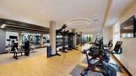 Located at 100 Chestnut St in Ashland, MA, this well-appointed fitness center offers a comprehensive exercise facility across a single, generously sized floor. The space is thoughtfully divided into distinct workout zones, each designed to support a variety of training needs.

The cardio area is lined with large windows that invite abundant natural light and offer views of the surrounding greenery, creating an energizing atmosphere. A row of treadmills and a stair climber are positioned to take full advantage of these outdoor views, while wall-mounted TVs provide additional entertainment. Recessed lighting and elegant circular pendant fixtures overhead complement the bright, airy feel of the space.

A dedicated cycling studio features a teal accent wall with vertical light strips and floor-to-ceiling mirrors, housing multiple stationary and air bikes for spin workouts. The strength training section is defined by dark, dramatic tones, with floor-length mirrors, linear LED accent lighting, and a full array of power racks, barbells, weight plates, and benches arranged on rubberized flooring.

The open main floor connects all zones seamlessly, finished in light hardwood-style flooring that lends warmth and continuity throughout. The overall ambiance is modern and professional, balancing high-performance functionality with a polished, welcoming aesthetic suited for a wide range of fitness routines.