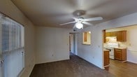 This single-floor space features an open living area finished with dark wall-to-wall carpeting and neutral white walls that create a clean, understated backdrop. A white ceiling fan with an integrated light fixture is centrally mounted, providing both air circulation and ambient lighting throughout the room. A textured ceiling adds subtle visual interest overhead.

Adjacent to the living area, a pass-through counter with a raised bar connects to the kitchen, which is outfitted with warm wood-toned cabinetry. This layout promotes an open flow between the living and kitchen spaces, making the area feel connected and functional. The kitchen can be partially viewed from the main living space, showcasing the cabinetry and countertop arrangement.

A hallway extends from the living area toward additional rooms, suggesting a practical floor plan with accessible private spaces beyond. Neutral-toned doors and trim throughout contribute to a cohesive interior aesthetic. HVAC venting is integrated into the ceiling, supporting climate control throughout the unit. The overall ambiance is straightforward and functional, offering a comfortable, move-in-ready environment with classic residential finishes.