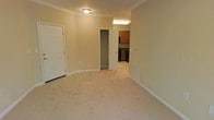 This single-floor residential unit offers a straightforward and functional layout designed for comfortable everyday living. The main living area features plush wall-to-wall carpeting in a neutral tone, complemented by warm beige walls and crisp white baseboards and crown molding that lend the space a clean, classic character. Flush ceiling lighting provides consistent illumination throughout. A built-in closet with a hanging rod is conveniently situated off the main living area, offering practical storage directly accessible from the central space. The entryway features tile flooring, providing a durable transition zone from the exterior. An in-wall vent is present along the baseboard, indicating a centralized climate system. The space flows directly into a compact kitchen, which is equipped with warm wood-toned upper and lower cabinetry, dark countertops, and black appliances. The kitchen benefits from overhead fluorescent lighting, ensuring a well-lit workspace. Crown molding carries throughout, maintaining a cohesive aesthetic across the unit. Overall, the space presents a tidy, neutral palette well-suited to a variety of furnishing styles, with a practical layout that maximizes usability within its footprint.