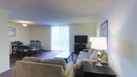 This single-floor space offers an open-concept layout that seamlessly connects the living and dining areas into one cohesive, functional environment. The living area is furnished with a generously sized sectional sofa in a neutral tone, paired with a rectangular coffee table that anchors the seating arrangement. A flat-screen television sits atop a low-profile media console positioned against the far wall, providing a straightforward entertainment setup. A stylish table lamp with a silver base adds a warm accent to the space. Large sliding glass doors fitted with vertical blinds allow an abundance of natural light to fill the room, creating a bright and airy atmosphere while offering access to the outdoors. The dining area, situated adjacent to the living space, features a compact table with dark wooden chairs, making it suitable for everyday use. Dark carpeting runs throughout, adding a layer of warmth underfoot. The walls are finished in soft, neutral tones that complement the overall understated aesthetic. The space is practical and well-organized, offering a comfortable living environment suited for everyday needs.