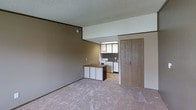 This single-floor space features an open layout connecting the living area to the kitchen. The living area is finished with wall-to-wall carpet in a neutral tone, complemented by brown accent walls and a textured popcorn ceiling. A transition from carpet to hard flooring marks the boundary between the living space and the kitchen area. The kitchen is equipped with white lower cabinetry and a peninsula that doubles as a workspace or casual dining counter with a dark laminate surface. Upper cabinetry with frosted or light-colored panel doors lines the kitchen walls, and a standard range is visible along the back wall. A sliding wood-paneled door is positioned near the kitchen entry, adding a warm, wood-toned element to the space. The overall aesthetic reflects a functional, utilitarian design with neutral tones throughout. The layout is practical and straightforward, making efficient use of the available floor space while maintaining an open feel between the main living and cooking areas.