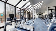 This contemporary fitness center is designed to inspire movement through an abundance of natural light and open, airy architecture. The space is defined by its striking sloped skylight ceiling and expansive floor-to-ceiling glass walls, which flood the interior with daylight and offer sweeping views of the surrounding landscape and outdoor terrace. The transparent enclosure creates a seamless connection between the indoor workout environment and the outdoors, making the space feel energizing and inviting at any time of day.

The cardio zone is prominently featured along the glass-lined perimeter, housing a row of modern treadmills and stationary bikes equipped with interactive digital screens for an engaging workout experience. The blue-toned carpet flooring adds a clean, purposeful aesthetic throughout the space while providing comfort underfoot.

Toward the interior, the fitness center transitions into a strength and functional training area, visible through the open layout. Ceiling fans and recessed lighting complement the natural light, ensuring the space remains comfortable and well-lit. The thoughtful layout allows for smooth circulation between equipment areas. Overall, this fitness center combines architectural elegance with practical functionality, offering members a motivating and well-equipped environment to pursue their fitness goals.