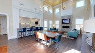 This single-floor open-concept space is designed to foster community and comfort in equal measure. The most striking architectural feature is the soaring double-height ceiling, finished with warm wood paneling and complemented by an elegant sculptural chandelier that anchors the central gathering area. A ceiling fan overhead ensures year-round comfort throughout the expansive layout.

The space is organized into distinct yet seamlessly connected zones. A kitchenette along one wall features white cabinetry, a tiled backsplash, and a bar-height counter lined with bold blue stools, ideal for casual dining or socializing. At the center of the room, a large farmhouse-style dining table is surrounded by a vibrant mix of teal, orange, and tan chairs, creating an inviting and eclectic atmosphere.

The lounge area is anchored by a floor-to-ceiling stacked stone fireplace with a wall-mounted television above, flanked by teal sofas and accent furniture arranged on a patterned area rug. Multiple windows flood the space with natural light, enhancing the bright, airy ambiance. Dark hardwood-style flooring runs throughout, grounding the space and adding warmth to the contemporary design.