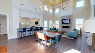 This single-floor open-concept space is designed to foster community and comfort in equal measure. The most striking architectural feature is the soaring double-height ceiling, finished with warm wood paneling and complemented by an elegant sculptural chandelier that anchors the central gathering area. A ceiling fan overhead ensures year-round comfort throughout the expansive layout.

The space is organized into distinct yet seamlessly connected zones. A kitchenette along one wall features white cabinetry, a tiled backsplash, and a bar-height counter lined with bold blue stools, ideal for casual dining or socializing. At the center of the room, a large farmhouse-style dining table is surrounded by a vibrant mix of teal, orange, and tan chairs, creating an inviting and eclectic atmosphere.

The lounge area is anchored by a floor-to-ceiling stacked stone fireplace with a wall-mounted television above, flanked by teal sofas and accent furniture arranged on a patterned area rug. Multiple windows flood the space with natural light, enhancing the bright, airy ambiance. Dark hardwood-style flooring runs throughout, grounding the space and adding warmth to the contemporary design.
