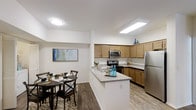 This single-story space showcases an open-concept layout that seamlessly connects the kitchen and dining area, creating a cohesive and functional living environment. The kitchen is outfitted with warm honey-toned wood cabinetry that extends to the upper corners, maximizing storage capacity. Granite-style countertops provide ample workspace and are complemented by a peninsula that doubles as a casual serving or prep area. Stainless steel appliances, including a refrigerator, electric range, and over-the-range microwave, lend a modern, polished look to the space. The flooring transitions from wood-look planks in the kitchen to a patterned area rug in the dining zone, subtly defining each area within the open floor plan. The dining area comfortably accommodates a round table and chairs, making it well-suited for everyday meals or casual gatherings. Overhead, recessed flat-panel lighting fixtures illuminate both the kitchen and dining space efficiently. White walls throughout keep the interior feeling bright and clean, while a piece of abstract wall art adds a subtle decorative touch. The overall ambiance is contemporary, practical, and welcoming.