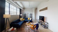 This single-floor unit showcases a striking architectural character defined by soaring vaulted ceilings and exposed structural beams and columns throughout, lending the space an industrial-loft aesthetic with considerable vertical volume. The open living area is generously proportioned, featuring large grid-pane windows that invite abundant natural light and views of the surrounding cityscape. Rounded architectural columns add visual interest and help delineate the living zone from a compact dining nook, which comfortably fits a small table and seating.

The kitchen is well-equipped with dark espresso cabinetry, stainless steel appliances including a refrigerator, range, microwave, and dishwasher, complemented by light countertops and a functional butcher-block island for additional workspace.

The primary bedroom features a dramatic vaulted ceiling, a walk-in closet, and a private en-suite bathroom with a shower. The second bedroom benefits from a wide band of industrial-style windows, filling the room with natural light. A second full bathroom with a shower-tub combination serves this room. A dedicated office or den space rounds out the layout, offering a quiet area suitable for work or study.