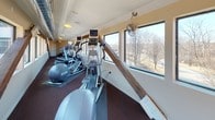 Located at 1 River Court in Amesbury, Massachusetts, this fitness center offers a well-appointed, single-floor exercise space designed to support an active lifestyle. The room is laid out in a long, corridor-style configuration, maximizing the available floor space while keeping all equipment easily accessible.

The facility is equipped with a comprehensive lineup of commercial-grade cardio machines, including treadmills, elliptical trainers, and a stationary recumbent bike, all from the Precor brand. Each machine is positioned to take full advantage of the expansive windows lining both sides of the space, allowing members to enjoy natural light and views of the surrounding neighborhood and treeline while working out.

A distinctive architectural element is the series of exposed, diagonal wooden support beams that run along the interior walls, adding a rustic, industrial character that contrasts with the otherwise clean, modern finishes. The ceiling features visible ductwork and recessed lighting, complementing the utilitarian yet comfortable aesthetic. Dark, durable carpeting covers the floor throughout, providing both cushioning and a polished look.

Ceiling-mounted fans enhance air circulation, contributing to a comfortable workout environment. The overall ambiance is bright, open, and functional — a practical fitness space that blends thoughtful design with quality equipment.