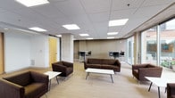 This single-floor space is thoughtfully designed as a versatile lounge and collaborative area, combining comfortable seating with functional workspaces. The open-plan layout is anchored by a recessed tile ceiling fitted with evenly spaced rectangular LED panel lights, ensuring consistent, bright illumination throughout. Light wood-toned flooring runs the length of the space, lending warmth and visual continuity.

The lounge area is furnished with dark brown upholstered sofas and armchairs arranged around low, white rectangular coffee tables, creating several inviting conversation groupings. A prominent exposed concrete column adds an industrial architectural element, contrasting with the otherwise refined interior. Along the perimeter, counter-height tables paired with black bar stools provide dedicated spots for focused work or casual dining.

One wall features a wall-mounted flat-screen television above a modest seating arrangement, offering a media-oriented zone within the space. A set of floor-to-ceiling glass sliding doors and expansive windows line an entire wall, flooding the interior with natural light and offering open views of a landscaped outdoor courtyard and surrounding buildings. A wood-framed interior door provides access to an adjacent room. The overall ambiance is modern, clean, and functional.