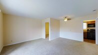 This single-floor space presents a clean, functional layout centered around a generously sized main room finished with wall-to-wall beige carpeting that lends a warm, understated feel underfoot. The walls are painted in a soft, neutral cream tone, contributing to a bright and airy atmosphere throughout. A ceiling fan with a light fixture is mounted centrally, providing both air circulation and ambient lighting to the room. The space features white baseboards that offer a crisp contrast against the light-colored walls and carpeting. A partial wall divider subtly defines a secondary area within the room, adding architectural interest while maintaining an open feel. An interior doorway leads to an adjoining room, hinting at additional usable space beyond the main area. The overall design is straightforward and move-in ready, with a neutral palette that offers flexibility for a variety of furnishing styles and uses. The single-floor layout ensures convenient, step-free access throughout the space, making it practical and accessible.