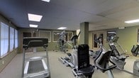 This fitness center offers a comprehensive workout environment designed to accommodate a range of exercise needs. The space is outfitted with an array of cardio machines, including ellipticals and a treadmill, as well as a multi-station cable weight machine for strength training. A recumbent exercise bike adds further versatility to the available equipment. The drop ceiling features evenly spaced recessed lighting panels that provide bright, consistent illumination throughout the room. Large mirrors line the walls, visually expanding the space and allowing users to monitor their form during workouts. The walls are finished in a muted olive-green tone, lending the room a calm, focused atmosphere. Carpeted flooring runs throughout, while a structural column near the center of the room adds an architectural element without disrupting the overall flow of the space. Framed panels on the walls contribute to a clean, finished aesthetic. An entryway with wood-framed glass doors is visible at the rear of the space, offering convenient access. Overall, the fitness center is thoughtfully laid out to maximize usability while maintaining a comfortable and organized environment.
