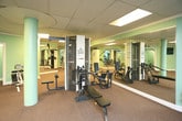 Located at 3901 Indian School Rd NE, this fitness center is a well-equipped, single-floor facility designed to support a comprehensive workout routine. The space is finished with neutral carpet flooring and soft mint-green walls, creating a clean and functional environment. A suspended drop ceiling with recessed panel lighting provides consistent, even illumination throughout the room.

The fitness center is outfitted with a variety of cardio machines, including treadmills, an elliptical trainer, and a stationary bike, alongside a range of strength-training equipment such as a Cybex cable machine, a weight bench, and a back extension bench. A central structural column anchors the layout of the room, naturally dividing the space into distinct workout zones. Full-length mirrors line multiple walls, visually expanding the space and allowing users to monitor their form during exercise.

A wall-mounted flat-screen television is positioned for easy viewing from the workout floor, adding an entertainment option during sessions. The room includes a secondary doorway leading to an adjoining area, as well as a waste bin and hand sanitizer station, reflecting attention to cleanliness and user convenience. The overall design is practical and straightforward, offering a fully functional fitness environment.