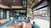 Located in Watertown, MA, this thoughtfully designed amenity space blends indoor comfort with outdoor living through a seamless, open-concept layout. The space is anchored by a sleek, modern kitchen featuring a dark-finished island with bar seating, white subway tile backsplash, open shelving, stainless steel appliances, and pendant lighting overhead. Adjacent dining areas with wood-topped tables and contemporary seating provide ample space for gathering and entertaining. Large-format floor-to-ceiling glass walls and black-framed sliding doors connect the interior directly to a spacious covered terrace, which is equipped with overhead infrared heaters for year-round use. The terrace features light stone tile flooring, exposed cream brick walls with a dark ceiling, and an inviting lounge area furnished with a generous sectional sofa in slate blue, colorful accent pillows, woven accent chairs, and a round coffee table set atop a warm-toned area rug. A separate outdoor patio section offers a wood-and-metal picnic table for al fresco dining, alongside additional seating and lush potted greenery. Retractable glass garage-style doors further blur the boundary between inside and out, flooding the space with natural light and open-air views of the surrounding Watertown landscape.