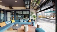 Located in Watertown, MA, this thoughtfully designed amenity space blends indoor comfort with outdoor living through a seamless, open-concept layout. The space is anchored by a sleek, modern kitchen featuring a dark-finished island with bar seating, white subway tile backsplash, open shelving, stainless steel appliances, and pendant lighting overhead. Adjacent dining areas with wood-topped tables and contemporary seating provide ample space for gathering and entertaining. Large-format floor-to-ceiling glass walls and black-framed sliding doors connect the interior directly to a spacious covered terrace, which is equipped with overhead infrared heaters for year-round use. The terrace features light stone tile flooring, exposed cream brick walls with a dark ceiling, and an inviting lounge area furnished with a generous sectional sofa in slate blue, colorful accent pillows, woven accent chairs, and a round coffee table set atop a warm-toned area rug. A separate outdoor patio section offers a wood-and-metal picnic table for al fresco dining, alongside additional seating and lush potted greenery. Retractable glass garage-style doors further blur the boundary between inside and out, flooding the space with natural light and open-air views of the surrounding Watertown landscape.