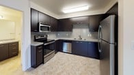This single-floor unit presents a clean, contemporary design throughout its well-organized layout. The kitchen is a standout feature, outfitted with dark navy cabinetry, granite-look countertops, and a full suite of stainless steel appliances including a refrigerator, electric range, built-in microwave, and dishwasher. A textured tile backsplash adds visual interest, while light-toned flooring ties the space together. Recessed fluorescent lighting illuminates the kitchen efficiently.

The open living area transitions seamlessly from the kitchen entry, featuring plush grey patterned carpeting and neutral white walls that create a bright, versatile canvas. A large closet with dark panel doors provides ample storage near the entry. The living space extends toward a glass door and window that open to a private balcony, allowing natural light to flow in and offering views of surrounding greenery.

The bedroom is a calm, well-proportioned space with the same grey carpeting, white walls, and a wide window fitted with horizontal blinds that frame outdoor tree views.

The bathroom features a full-size soaking tub with an overhead shower, a curved curtain rod, brushed nickel fixtures, tile flooring, and a granite-topped vanity — delivering a functional and polished finish.