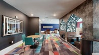 Located at 7222 S Peek Rd in Houston, Texas, this single-floor communal lounge delivers a dynamic, contemporary design that balances functionality with bold visual character. The space is unified by a striking multicolored geometric carpet that anchors the entire floor, providing a lively foundation for the varied seating arrangements throughout.

The layout is thoughtfully divided into multiple functional zones. A casual dining and gathering area features round wooden tables paired with teal upholstered chairs, positioned beneath recessed lighting against deep charcoal-painted walls. An adjacent lounge zone offers a curved rust-orange sofa with a low side table, set against a vivid cobalt blue accent wall. Privacy-focused seating booths with high cream-colored partitions provide semi-enclosed areas ideal for focused work or quiet conversation.

A dedicated workstation area lines one wall, equipped with desktop computers on a long floating counter with bar-height seating, illuminated by natural light from large windows that frame outdoor views. A textured feature wall with an architectural teal laser-cut panel design serves as a prominent focal point, adding artistic depth. Additional seating alcoves with booth-style benches and red accent walls contribute further variety.

The space is illuminated throughout by recessed ceiling lights and natural light, creating a bright and energetic atmosphere well-suited for collaboration, work, or relaxation.