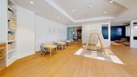 Located at 1600 Harbor Blvd in Weehawken, NJ, this thoughtfully designed amenity space spans a generous single floor, offering a diverse array of gathering and activity areas. The open-concept layout is anchored by a sleek, contemporary kitchen and bar area outfitted with expansive white quartz countertops, dark cabinetry, stainless steel appliances, and striking copper pendant lighting that adds warmth and sophistication. Long bar-height counters with modern stools create an inviting setting for casual socializing or communal dining. Adjacent to the kitchen, a comfortable lounge area features plush blue modular seating, layered lighting with oversized circular fixtures, and large windows that flood the space with natural light and views of the surrounding greenery. A more intimate seating nook showcases a deep teal accent wall, decorative mirrors, a wall-mounted display, and curved upholstered chairs arranged around a central coffee table, ideal for quiet conversation. A dedicated children's area is thoughtfully appointed with warm hardwood flooring, child-sized tables and chairs, a wooden climbing structure, foam play mats, and a star-themed media nook with low-slung seating. A utility room, closet, and bathroom round out the practical amenities of this well-appointed community space.
