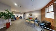 Located at 1630 Aliceanna St in Baltimore's vibrant Fells Point neighborhood, this single-floor amenity space serves as both a welcoming lounge and a functional business center. The layout is divided into two distinct zones that work together cohesively. The lounge area features a comfortable seating arrangement with a patterned sofa accented by colorful throw pillows, sleek leather armchairs, and a rustic wood coffee table — all centered on plush, patterned carpeting. A striking exposed brick wall anchors this section, lending warmth and industrial character, while windows allow natural light to filter in. Wall art adds a local Baltimore touch to the relaxed atmosphere. The adjacent business center is equipped with a built-in countertop workstation featuring two desktop computers, a printer, and warm wood cabinetry below — ideal for remote work or administrative tasks. Pendant lighting illuminates the workspace, creating a focused yet inviting environment. A potted indoor tree near the connecting corridor adds a refreshing natural element. Recessed ceiling lighting throughout ensures consistent, even illumination across the entire space. A restroom is conveniently accessible, and glass-paneled entry doors connect the space to the broader building corridor, enhancing accessibility and openness.