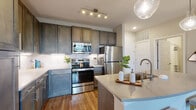 This thoughtfully designed single-floor residence offers a cohesive open-concept layout connecting the kitchen, dining, and living areas. The kitchen is a standout feature, showcasing rich dark-toned cabinetry, stainless steel appliances including a refrigerator, range, and built-in microwave, complemented by a large central island with a quartz countertop, undermount sink, and seating for two. Stylish pendant lighting hangs above the island, adding a modern touch. Warm hardwood-style flooring runs throughout the main living areas, creating a seamless flow.

The living room is anchored by a bold teal accent wall, built-in shelving, and large windows that invite natural light. The space is further enhanced by a ceiling fan and tasteful decor that adds personality and warmth.

The primary bedroom features plush carpeting, natural light from well-dressed windows, a ceiling fan, and access to a generously sized walk-in closet with wire shelving and ample hanging space. A second bedroom offers similar comfort and natural light. The bathroom provides a full tub-shower combination and tile flooring.

A connecting hallway with dark cabinetry and tile flooring links the bathroom and kitchen, contributing to a well-organized and functional floor plan throughout.
