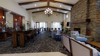 This single-floor communal space is defined by its striking vaulted ceiling adorned with dark-stained exposed wood beams, lending the interior a lodge-inspired character. An elegant chandelier hangs as a central focal point, complementing the recessed lighting throughout. The generous ceiling height enhances the open, airy feel of the room.

The layout is thoughtfully divided into functional zones. A bar-height counter with leather stools lines one side, providing a casual seating area ideal for socializing. Adjacent to it, a dining area features standard-height tables and chairs arranged near large windows that allow ample natural light to fill the space. The lounge area centers around comfortable upholstered seating arranged around a prominent stacked-stone fireplace, which anchors the room with warmth and texture. A wall-mounted television above the fireplace adds a modern amenity.

Patterned broadloom carpet with an abstract design covers much of the floor, adding visual interest underfoot. Decorative artwork, potted plants, and warm wall sconces contribute to the inviting, upscale ambiance throughout the space.
