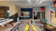 The Club Room at 290 George St in New Brunswick, NJ is a thoughtfully designed communal amenity space that blends modern aesthetics with a welcoming, energetic atmosphere. The open-plan layout accommodates a variety of social settings, anchored by a generous sectional sofa accented with vibrant yellow and teal throw pillows, set atop a patterned area rug. A striking bar-height counter with a granite surface and deep teal cabinetry serves as a central gathering point, complemented by matching teal barstools and pendant lighting overhead. Teal accent chairs with bold orange frames are arranged throughout for intimate conversation areas. A standout feature is the large-scale, colorful mural that adds artistic energy to one wall, while a reclaimed wood accent wall adorned with sculptural golden leaf décor creates a warm, textural contrast. A built-in window seat with a teal cushion and natural light from large windows offers a quiet retreat within the space. Linear LED ceiling fixtures cast a modern, well-lit ambiance throughout. Large interior windows provide views into a model kitchen, offering residents a preview of available unit finishes. The overall design is cohesive, contemporary, and vibrant — ideal for socializing, relaxing, or casual gatherings.