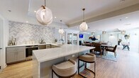This single-floor space offers a seamlessly connected open-plan layout encompassing a kitchen, dining room, and living room, all unified by warm hardwood flooring and crisp white crown molding throughout. The kitchen serves as a central showpiece, anchored by a generous island topped with striking veined white marble-style quartz and surrounded by upholstered bar stools. Gray shaker cabinetry, a herringbone tile backsplash, and globe pendant lights in brushed brass lend the space a refined yet approachable aesthetic. Stainless steel appliances and a deep-basin sink complete the highly functional workspace. Adjacent to the kitchen, the dining area accommodates a substantial walnut dining table paired with upholstered chairs, all set atop a patterned area rug. French doors with glass panes connect the dining room to the living area, offering both openness and definition. The living room is a standout, featuring a floor-to-ceiling stacked stone fireplace with a wall-mounted TV above, flanked by built-in wood-paneled bench seating with cushions. Statement gold chandelier fixtures with cascading metallic fronds add dramatic flair, while large windows invite natural light and outdoor views. The overall ambiance is sophisticated, warm, and cohesive.