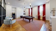 Located at 2110 Pickens St in Columbia, SC, this single-floor amenity space spans an impressive footprint and offers a thoughtfully designed environment that blends historic character with modern finishes. The wide, light-filled corridors serve as the connective tissue of the space, featuring rich hardwood floors, globe pendant lighting, and walls finished in deep, sophisticated tones of forest green and navy — lending a polished, cohesive aesthetic throughout. Steel-framed glass partitions provide visual transparency between areas while maintaining distinct zones, adding an industrial-modern architectural element to the design. The fitness studio is a standout feature, offering durable speckled flooring, large windows for natural light, ceiling fans, wall-mounted mirrors, a ballet barre, stationary bikes, and a full rack of exercise equipment. Adjacent to the fitness area, the game room provides an inviting social space complete with hardwood floors, a full-size pool table, arcade machine, built-in shelving, a wall-mounted display, and natural light streaming through tall windows dressed in bold red curtains. Throughout the space, globe and multi-bulb pendant fixtures contribute a warm, contemporary ambiance, while the generous proportions of each area create an open, welcoming atmosphere well-suited for community and recreation.