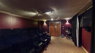 This dedicated movie screening room is designed to deliver an authentic cinematic experience. The room is dressed in deep crimson walls that set a moody, theater-like atmosphere, complemented by patterned carpeting that adds a classic cinema aesthetic throughout the space. Track lighting mounted along the textured ceiling provides adjustable ambient illumination, while wall-mounted sconces offer warm accent lighting along the sides.

Seating is arranged in a tiered, stadium-style layout with multiple rows of black cushioned theater chairs, each equipped with armrests and cup holders for comfort during screenings. The raised rear platform ensures unobstructed sightlines from every seat in the room. A large wall-mounted flat-screen display serves as the primary viewing screen, framed by floor-length black blackout curtains that run the length of the front wall, effectively eliminating light interference for an immersive viewing environment.

Near the entrance, a classic popcorn machine adds a fun, concession-stand touch, further enhancing the movie theater experience. The solid wood entry door provides a clean separation from adjoining spaces. Overall, this screening room is thoughtfully appointed to provide a comfortable, fully immersive movie-watching environment suitable for private screenings or group gatherings.