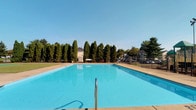 Located at 5280 Tamarack Cir E in Columbus, Ohio, this single-level outdoor amenity space centers around a generously sized community swimming pool. The pool features crystal-clear blue water and is framed by a wide concrete deck that provides ample space for lounging and movement around the perimeter. Sturdy metal handrails offer safe and convenient entry into the water.

The area is enclosed by wrought iron fencing, providing both security and an open, airy feel. A striking row of tall, columnar evergreen trees lines the back boundary of the space, creating a natural privacy screen and lending a polished, manicured appearance to the setting. Additional mature deciduous trees are visible beyond the fence line, contributing to the lush, green atmosphere of the surroundings.

The open-sky setting allows for abundant natural light throughout the day, making this an inviting destination during warm months. Neighboring structures are visible in the background, placing this amenity within a residential community context. The overall ambiance is clean, well-maintained, and community-oriented, offering a refreshing outdoor retreat for residents of the Tamarack Circle area of Columbus.
