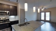 This single-floor residence showcases a thoughtfully designed open-concept layout that seamlessly connects the kitchen and living areas. The kitchen is a standout feature, anchored by a spacious island topped with granite countertops and equipped with a double stainless steel sink and a sleek gooseneck faucet. Three cylindrical pendant lights hang above the island, providing both functional task lighting and a modern aesthetic touch. Dark espresso-toned cabinetry lines the walls, offering generous storage while creating a striking contrast against the neutral grey walls and light-toned granite surfaces. A classic tile backsplash adds texture and detail behind the countertops. The adjacent living area benefits from a ceiling fan and access to natural light through a windowed door, suggesting a connection to an outdoor space. The overall ambiance is clean, contemporary, and well-appointed, with consistent finishes throughout that reflect a cohesive design vision. The open flow between spaces makes the interior feel airy and functional, ideal for everyday living and entertaining alike.