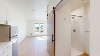 Located at 4612 Stone Way N in Seattle's vibrant Wallingford neighborhood, this thoughtfully designed studio makes efficient use of its single-floor layout. The main living area is bright and airy, featuring light hardwood flooring, crisp white walls, and large windows on two sides that flood the space with natural light and offer pleasant views of the surrounding neighborhood under Seattle's open sky.

The kitchenette is efficiently outfitted with white cabinetry, a stainless steel refrigerator, an electric cooktop, a microwave, an integrated sink, and an in-unit washer — all neatly arranged along one wall. A bold red entry door adds a striking pop of color and serves as a design focal point in the kitchen area.

The bathroom is accessed through a stylish sliding barn door with a dark metal handle, lending a modern industrial touch to the interior. Inside, the bathroom features a walk-in shower with a rainfall showerhead, a clean white vanity, and warm wood-tone flooring, all finished with minimalist chrome fixtures and accessibility grab bars.

Recessed lighting and flush-mount ceiling fixtures provide consistent illumination throughout. The overall design aesthetic is clean, modern, and functional — well-suited for urban living in one of Seattle's most walkable and lively neighborhoods.