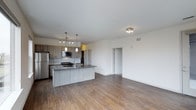 This single-floor space showcases a thoughtfully designed open-concept layout that seamlessly blends the kitchen and living areas into one cohesive, airy environment. The kitchen is a standout feature, anchored by a substantial central island with a dark countertop, an integrated sink, and seating potential. Cabinetry in a warm taupe wood finish lines the walls, complemented by a classic subway tile backsplash and a full suite of stainless steel appliances, including a refrigerator, microwave, and gas range. Pendant lighting above the island adds both function and contemporary style. The open living area extends generously from the kitchen, bathed in natural light and finished with warm-toned, hardwood-style flooring that runs throughout. Crisp white walls and a flush ceiling light fitting keep the space feeling clean and bright. A doorway visible from the main area leads to additional rooms, hinting at further functional spaces beyond. The overall ambiance is modern and inviting, with a neutral palette that offers a versatile backdrop suited to a wide range of interior styles.