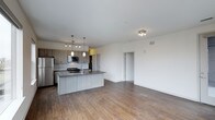 This single-floor space showcases a thoughtfully designed open-concept layout that seamlessly blends the kitchen and living areas into one cohesive, airy environment. The kitchen is a standout feature, anchored by a substantial central island with a dark countertop, an integrated sink, and seating potential. Cabinetry in a warm taupe wood finish lines the walls, complemented by a classic subway tile backsplash and a full suite of stainless steel appliances, including a refrigerator, microwave, and gas range. Pendant lighting above the island adds both function and contemporary style. The open living area extends generously from the kitchen, bathed in natural light and finished with warm-toned, hardwood-style flooring that runs throughout. Crisp white walls and a flush ceiling light fitting keep the space feeling clean and bright. A doorway visible from the main area leads to additional rooms, hinting at further functional spaces beyond. The overall ambiance is modern and inviting, with a neutral palette that offers a versatile backdrop suited to a wide range of interior styles.
