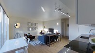 This single-floor residence features an open-concept layout that integrates the living, dining, and kitchen areas into one cohesive space. The living area is anchored by a navy blue sectional sofa positioned atop a geometric patterned area rug, complemented by a pair of dark leather accent chairs. A flat-screen television sits across from the seating arrangement, while a sleek industrial-style console table with metal X-frame detailing serves as a functional centerpiece.

Overhead, a track lighting system runs along the ceiling, providing directed illumination throughout the space. The neutral, light-toned walls and smooth white ceiling enhance the sense of openness and brightness. Warm-toned hardwood flooring extends throughout, adding a natural, cohesive element to the interior.

A wall-mounted gallery arrangement of six framed pieces adds visual interest near the living area. A floor lamp and wall sconce provide additional ambient lighting options. The dining area features a white table and chairs near the perimeter. A hallway visible in the background leads to additional rooms, suggesting a practical and well-organized floor plan throughout.