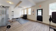 This single-floor fitness center is designed to support a well-rounded workout routine within a bright, clean environment. The space is divided into distinct functional zones, each tailored to different types of exercise. The entry area transitions from tile flooring into carpeted workout zones, providing a practical layout that separates high-traffic pathways from active training areas.

The strength training zone is well-appointed, featuring a multi-tier dumbbell rack stocked with a full range of weights, a weight bench, a cable machine, a barbell with plates, and an inversion table. Protective rubber and interlocking floor mats cover the workout areas, offering cushioning and stability during exercise. A ceiling fan overhead helps maintain air circulation throughout the space.

Natural light filters in through multiple double-hung windows along the exterior wall, offering views of the surrounding greenery and keeping the atmosphere open and inviting. The cardio area accommodates a treadmill and additional equipment, rounding out the facility's offerings.

Neutral-toned walls, consistent overhead lighting, and straightforward access through a dedicated exterior door contribute to the center's functional and organized character, making it a practical resource for daily fitness needs.