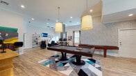 This expansive game room is designed to offer a well-rounded recreational experience within a single, open-plan layout. The space features wood-look flooring throughout, high ceilings with crown molding, and recessed lighting complemented by large pendant fixtures that hang above the central pool table — a dark-finished centerpiece set atop a geometric area rug in teal, gray, and white tones.

Adjacent to the pool table sits a shuffleboard table, providing additional gaming options. A dedicated computer bar station with bar-height seating and a dark textured surround offers a functional workspace. Nearby, a framed oversized Scrabble board serves as both a decorative and interactive feature mounted on the wall.

The lounge area includes black swivel armchairs arranged around open metal shelving units displaying decorative accessories, with a wall-mounted flat-screen TV as a focal point. Natural light enters through multiple windows dressed with curtain panels, brightening the white walls.

A wet bar or refreshment counter with white cabinetry, granite countertops, and a bold geometric-patterned accent wall adds a stylish, functional touch. The overall ambiance is modern and inviting, blending entertainment, comfort, and contemporary design seamlessly throughout the room.