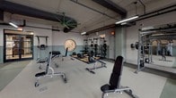 This expansive fitness center is organized into several distinct workout zones, each thoughtfully designed to support a comprehensive exercise experience. The main cardio area features a row of modern treadmills and stationary bikes positioned directly in front of floor-to-ceiling windows, offering unobstructed views of the surrounding landscape and abundant natural light. Additional cardio equipment, including rowing machines and stair climbers, occupies a separate section with an exposed ceiling showcasing industrial-style ductwork and pipe installations illuminated by linear pendant lighting.

The strength training area is outfitted with power racks, barbells, weight plates, and adjustable benches arranged on durable rubberized flooring. Large mirrors line the walls, enhancing the sense of space. A dedicated cycling studio features warm hardwood flooring, dark green wainscoting, pendant lighting, and direct outdoor access through glass doors. An adjoining functional fitness room with floor-to-ceiling mirrors and a glass partition wall houses medicine balls and additional training equipment. A large green industrial ceiling fan adds a distinctive visual element throughout the main workout floor, complementing the facility's modern yet utilitarian aesthetic.