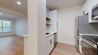 This single-floor residence showcases a clean, contemporary design highlighted by light hardwood flooring that runs consistently throughout the space, creating a cohesive and airy aesthetic. The open-concept living area is bright and welcoming, benefiting from a large picture window that frames lush outdoor greenery and allows natural light to fill the room. A built-in shelving unit along one wall provides both functional storage and a subtle architectural detail.

The galley-style kitchen is efficiently designed with white shaker cabinetry featuring matte black hardware, white quartz countertops, and a marble-style tile backsplash. Open wood floating shelves add warmth and contrast against the cabinetry. Stainless steel appliances, including a refrigerator, electric range, over-the-range microwave, and dishwasher, complete the kitchen's modern, functional layout.

The bathroom features a soaking tub and shower combination enclosed by a frameless glass panel, finished with large-format white marble-look tile and matte black fixtures. A conveniently adjacent laundry closet with a full-size washer adds practical utility. The bedrooms are bright and well-proportioned, each featuring hardwood flooring, neutral walls, and windows with outdoor views.