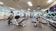 Located at 1121 W Prospect Rd in Fort Collins, Colorado, this fitness center offers a spacious, single-floor layout designed to accommodate a wide variety of workout routines. The facility is characterized by its drop-panel ceiling fitted with recessed fluorescent lighting, ceiling fans for air circulation, and durable rubber flooring throughout — all practical elements that contribute to a clean, functional environment.

Natural light is a notable feature, with rows of horizontal windows lining multiple walls and a glass door providing views of the surrounding tree-lined exterior. Large mirrors are strategically placed to enhance the sense of space and allow users to monitor their form during exercise.

The cardio section is well-appointed, housing multiple treadmills, ellipticals, stationary bikes, and stair climbers, many equipped with individual screens for entertainment. The strength training area includes a broad selection of cable machines, multi-station weight equipment, adjustable benches, a full dumbbell rack, barbells, and stability balls, catering to both beginner and experienced users. Mounted flat-screen televisions are positioned throughout the space for added convenience. The overall atmosphere is bright, organized, and purpose-built for fitness.