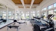 Located in Goodyear, Arizona, this single-floor fitness center combines elegant architectural design with a comprehensive selection of exercise equipment. The space is defined by its soaring coffered ceilings adorned with warm wood paneling and structural beams, lending an upscale aesthetic rarely found in community fitness facilities. Large arched windows line the walls, flooding the interior with abundant natural light and offering views of the surrounding landscaped grounds, including mature trees and palm-lined walkways. The light-toned herringbone-patterned flooring enhances the bright, airy atmosphere throughout. Full-length mirrors along one wall expand the sense of space and allow users to monitor their form during workouts. Equipment is thoughtfully distributed across the open floor plan and includes multiple treadmills with integrated screens, elliptical trainers, a stationary bike, rowing machines, a cable strength training system, adjustable benches, and a dedicated dumbbell rack. Ceiling fans provide additional comfort during workouts. A glass-paneled entry door beneath an arched surround connects the space to the exterior, allowing for seamless indoor-outdoor flow. The overall ambiance is clean, modern, and welcoming, making this fitness center a functional and visually refined space for daily exercise.