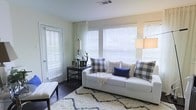This single-floor space features a welcoming living area designed with a clean, modern farmhouse aesthetic. The living room is bathed in natural light, courtesy of multiple windows fitted with white horizontal blinds and sheer white curtains that soften the incoming daylight. The walls are painted in a crisp white, contributing to the bright and airy atmosphere throughout the room. Dark hardwood flooring anchors the space, providing a rich contrast to the lighter elements and adding warmth to the overall design. A plush, white area rug with a subtle geometric pattern defines the seating area, adding texture and comfort underfoot. The centerpiece of the room is a white sofa adorned with an assortment of buffalo check and plaid throw pillows in grey, white, and navy tones, complemented by a cozy woven throw blanket. A sleek dark side table holds a classic table lamp and decorative accents, while a bench seat near the entry adds functional seating. The space also benefits from a glass-paneled door that introduces additional light. Overall, the living area exudes a relaxed yet polished ambiance, well-suited for comfortable everyday living.