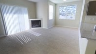 This single-floor space offers a clean, neutral aesthetic throughout. The living area serves as a central highlight, featuring wall-to-wall carpeting in a warm beige tone that adds softness underfoot and contributes to the overall sense of comfort. A white-mantled fireplace anchors one corner of the room, providing a classic focal point and a cozy atmosphere. Natural light streams generously through multiple windows, including a large multi-pane window that frames an outdoor view of neighboring rooftops and mature trees. Vertical blinds on an adjacent window offer flexible light control. The walls are finished in a soft, neutral gray-beige palette that complements the light carpeting and creates a calm, inviting ambiance throughout. Clean white baseboards provide a crisp contrast to the walls, and the ceiling-mounted light fixture adds practical overhead illumination. The open layout of the living area allows for versatile furniture arrangements, making the space well-suited for a variety of uses. Overall, the space presents a simple, functional design with timeless finishes and a bright, airy feel that makes it immediately welcoming.