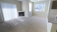 This single-floor space offers a clean, neutral aesthetic throughout. The living area serves as a central highlight, featuring wall-to-wall carpeting in a warm beige tone that adds softness underfoot and contributes to the overall sense of comfort. A white-mantled fireplace anchors one corner of the room, providing a classic focal point and a cozy atmosphere. Natural light streams generously through multiple windows, including a large multi-pane window that frames an outdoor view of neighboring rooftops and mature trees. Vertical blinds on an adjacent window offer flexible light control. The walls are finished in a soft, neutral gray-beige palette that complements the light carpeting and creates a calm, inviting ambiance throughout. Clean white baseboards provide a crisp contrast to the walls, and the ceiling-mounted light fixture adds practical overhead illumination. The open layout of the living area allows for versatile furniture arrangements, making the space well-suited for a variety of uses. Overall, the space presents a simple, functional design with timeless finishes and a bright, airy feel that makes it immediately welcoming.
