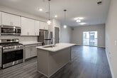 Located at 36 S New York Rd in Galloway, NJ, this single-floor unit showcases a clean, contemporary design with consistent wood-style flooring and a neutral warm-beige color palette throughout. The open-concept layout connects the kitchen and living area seamlessly, creating a spacious and functional flow.

The kitchen is well-appointed with white upper cabinetry, gray lower cabinetry, and a full suite of stainless steel appliances, including a gas range, microwave, and refrigerator. A central island with a white quartz countertop and undermount sink serves as both a prep area and a visual anchor for the space, accented by pendant lighting above.

The living area is generously proportioned, benefiting from natural light and direct access to a private balcony through sliding glass doors, extending the living space outdoors. The bedroom offers natural light through a window with blinds, a walk-in closet with built-in shelving, and a connecting door to adjacent spaces.

The bathroom features a tiled tub-shower combination with a striking contrast of white marble-style tile and dark accent tile, complemented by marble-look flooring, providing a polished, modern aesthetic throughout.