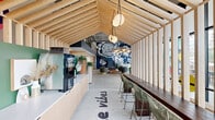 Located at 914 S Milton Rd in Flagstaff, Arizona, this dynamic lobby and common area serves as the social heart of the Yugo Flagstaff community. The single-floor space is generously sized, featuring soaring double-height ceilings with exposed concrete and industrial-style ductwork that lend a contemporary, urban feel. A striking black-and-white topographic mural dominates one wall, complemented by a bold navy-blue color palette that gives the space a distinctive, energetic character.

Floor-to-ceiling windows along the street-facing facade flood the interior with abundant natural light while offering open views of the surrounding Flagstaff streetscape. Colorful sculptural elements hang in the window bay, adding an artistic touch. The main entrance leads into an expansive open-plan area with large-format tile flooring, furnished with curved lounge chairs and casual seating clusters ideal for socializing.

A recreation zone includes a ping-pong table and foosball table, encouraging community engagement. A sleek reception desk with warm wood slatted accents and the Yugo branded signage anchors the service area, alongside resident mailboxes and access to building corridors. A metal-and-wood staircase with cable railings connects to an open mezzanine level above, further enhancing the sense of vertical space. Statement pendant lighting throughout adds warmth and visual interest to this well-designed, community-focused space.