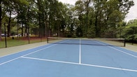 This athletic facility features a full-size outdoor tennis court surfaced in a two-tone blue and green hard court finish, with clearly marked white lines and a regulation net at center. The court is fully enclosed by black chain-link fencing, providing a secure and defined playing area. Towering mature trees surround the perimeter, creating a natural, wooded backdrop that offers shade and a sense of privacy. A brick structure adjoins the court on one side, housing a dedicated fitness room that connects directly to the outdoor space via a glass-panel door. The fitness room features rubber speckled flooring suited for exercise equipment, gray-painted walls, and a textured ceiling. Natural light enters through several windows, maintaining views of the surrounding greenery and the court beyond. The room accommodates cardio machines, including a treadmill and elliptical, as well as a multi-station strength training system. A wall-mounted television and a corkboard are also present. Together, the tennis court and fitness room form a cohesive, self-contained athletic complex designed for both outdoor play and indoor training.
